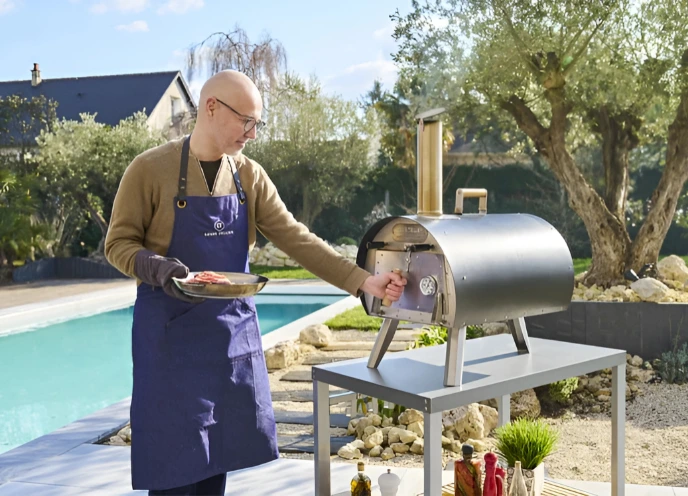 Homme en tablier Louis Tellier utilisant le four à bois extérieur Marcel dans un jardin près d'une piscine, prêt à enfourner des aliments