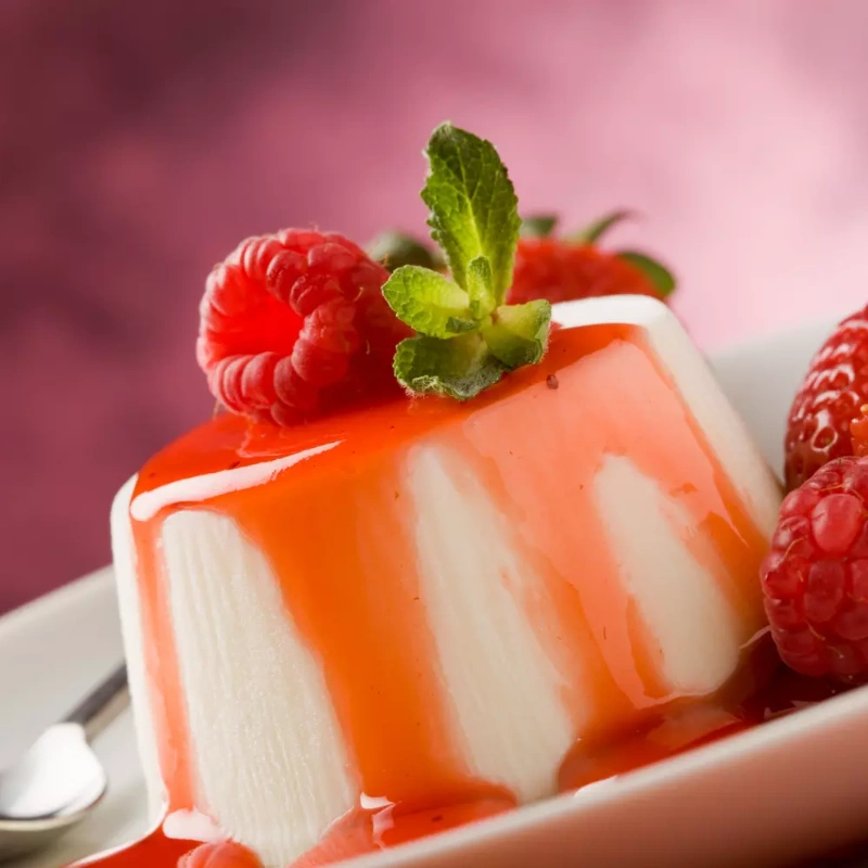 Panna cotta nappée de coulis rouge, accompagnée de framboises et menthe fraîche